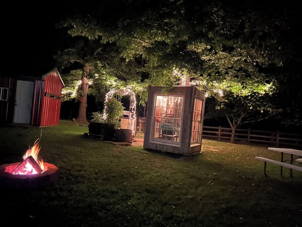 Une scène nocturne en plein air, avec un foyer, une structure en bois illuminée et un bâtiment rouge en arrière-plan.