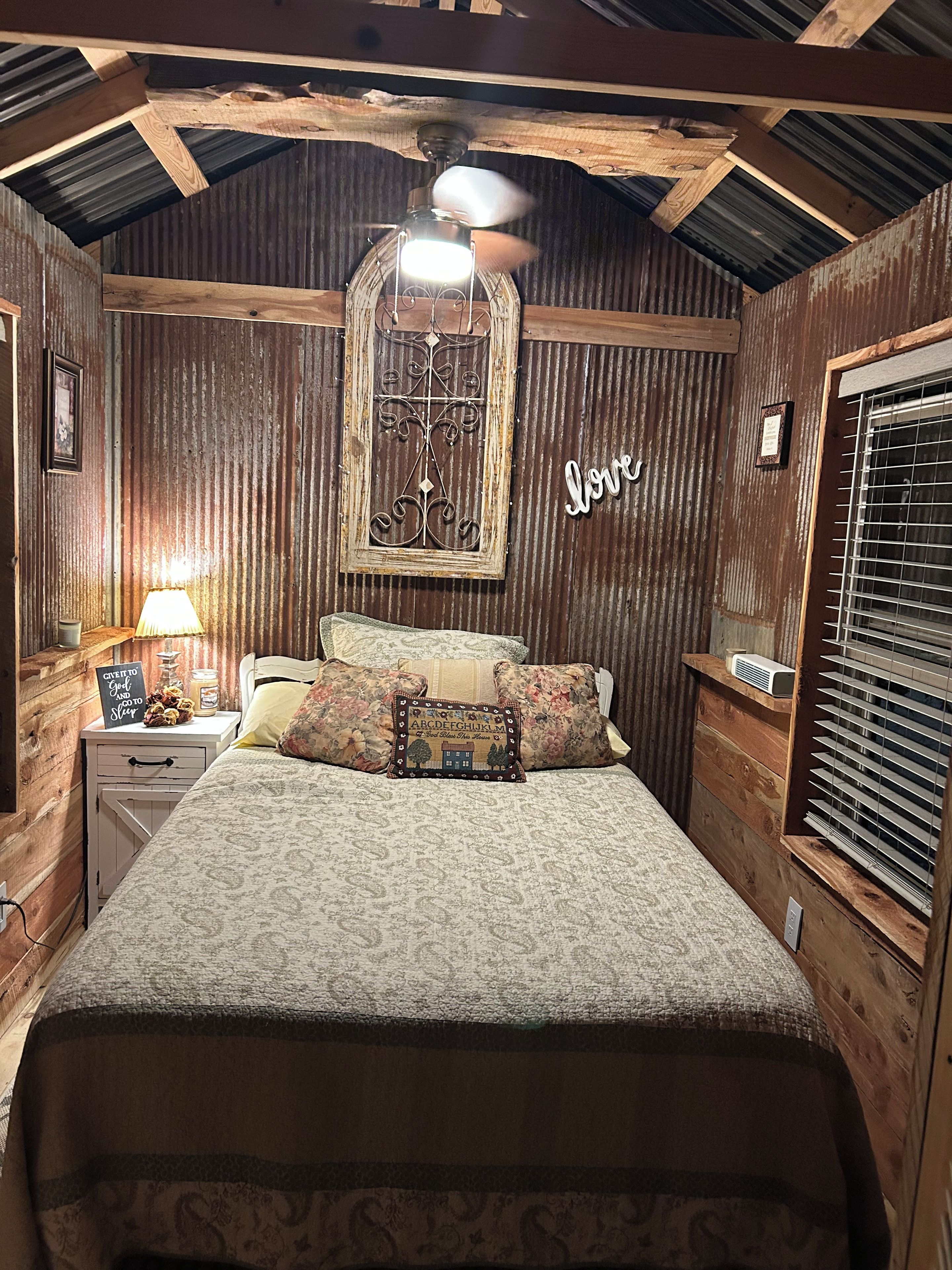Un dormitorio con techo de madera, una cama con ropa de cama floral, una mesita de noche con una lámpara y una ventana con persianas.