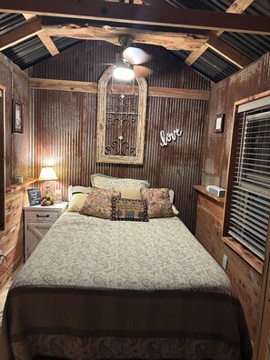 Une chambre avec un plafond en bois, un lit recouvert d’une literie à motifs floraux, une table de chevet avec une lampe et une fenêtre avec des stores.
