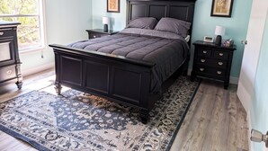 A bedroom with a dark wooden bed, a large window with blinds, a ceiling fan, and a patterned rug. - 3-bedroom Private Forest Retreat Home with Wifi and Full Kitchen (Spring Hill)