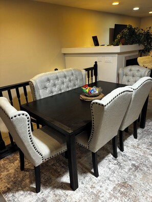 A dining room with a dark wooden table surrounded by six upholstered chairs with button-tufted backs. A woven placemat holds a bowl of snacks.
