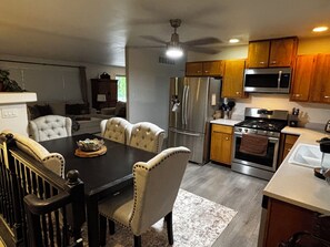 A modern kitchen with stainless steel appliances, a dining area with a dark wood table and upholstered chairs, and a ceiling fan.