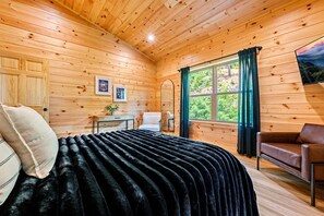 A cozy wooden cabin room with a bed, a chair, a table, and a window with curtains.