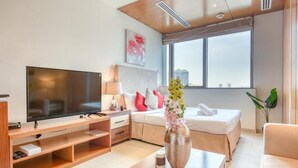 Ein modernes Hotelzimmer mit Flachbildfernseher, einem Holztischchen, einem Bett mit weißer Bettwäsche, einem Nachttisch mit Lampe und einem Fenster mit Blick auf die Stadt.
