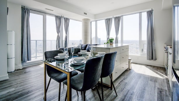 A modern dining room with a glass table, dark chairs, and a view of the city through large windows.