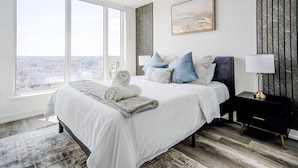 A modern bedroom with a large bed, a nightstand, a lamp, and a city view.