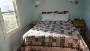 A bedroom with a quilted bedspread, two bedside lamps, a nightstand, and a window with a view of a house and lawn.