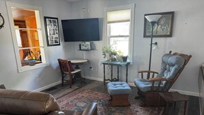 A living room with a brown leather sofa, a blue armchair, a small table with a plant, a television mounted on the wall, and a large mirror.