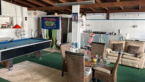 A pool table, wicker chairs, a glass-top table with a vase, and a couch in a room with a high ceiling and exposed beams.
