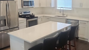 A modern kitchen with white cabinetry, stainless steel appliances, a central island with bar stools, and a window with blinds.