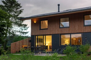 Una casa moderna de dos pisos con fachada de madera, ventanas grandes y un patio con mobiliario exterior.