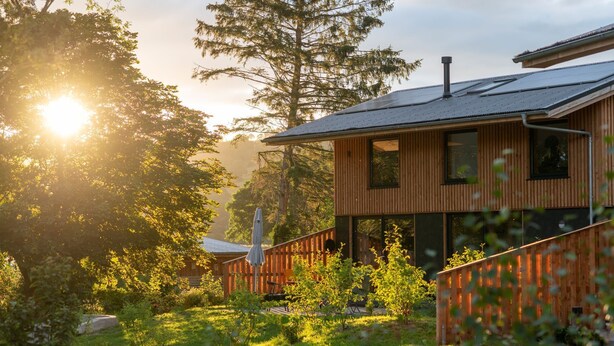 A modern wooden cabin with a sloped roof, surrounded by lush greenery and a clear path leading to the entrance.