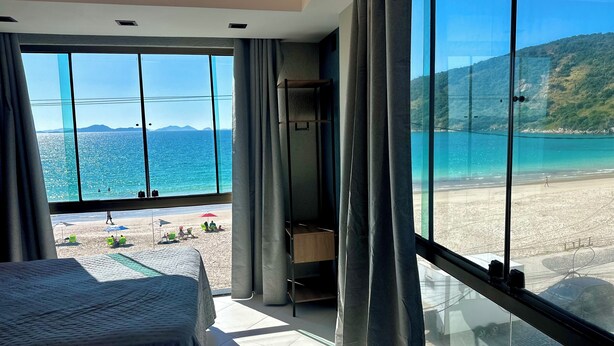 A room with a large window overlooking a beach, a bed, and a small seating area. - LOFT JANELAS DO CARIBE - 315 (Arraial do Cabo)