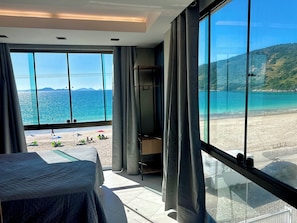 Una habitación con un ventanal grande con vistas a la playa, una cama y una pequeña zona de estar.
