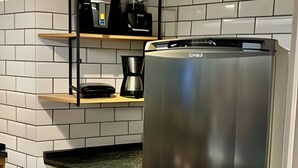 A kitchen with white tiled walls, a stainless steel refrigerator, a coffee maker, and a shelf with a kettle. - LOFT JANELAS DO CARIBE - 315 (Arraial do Cabo)