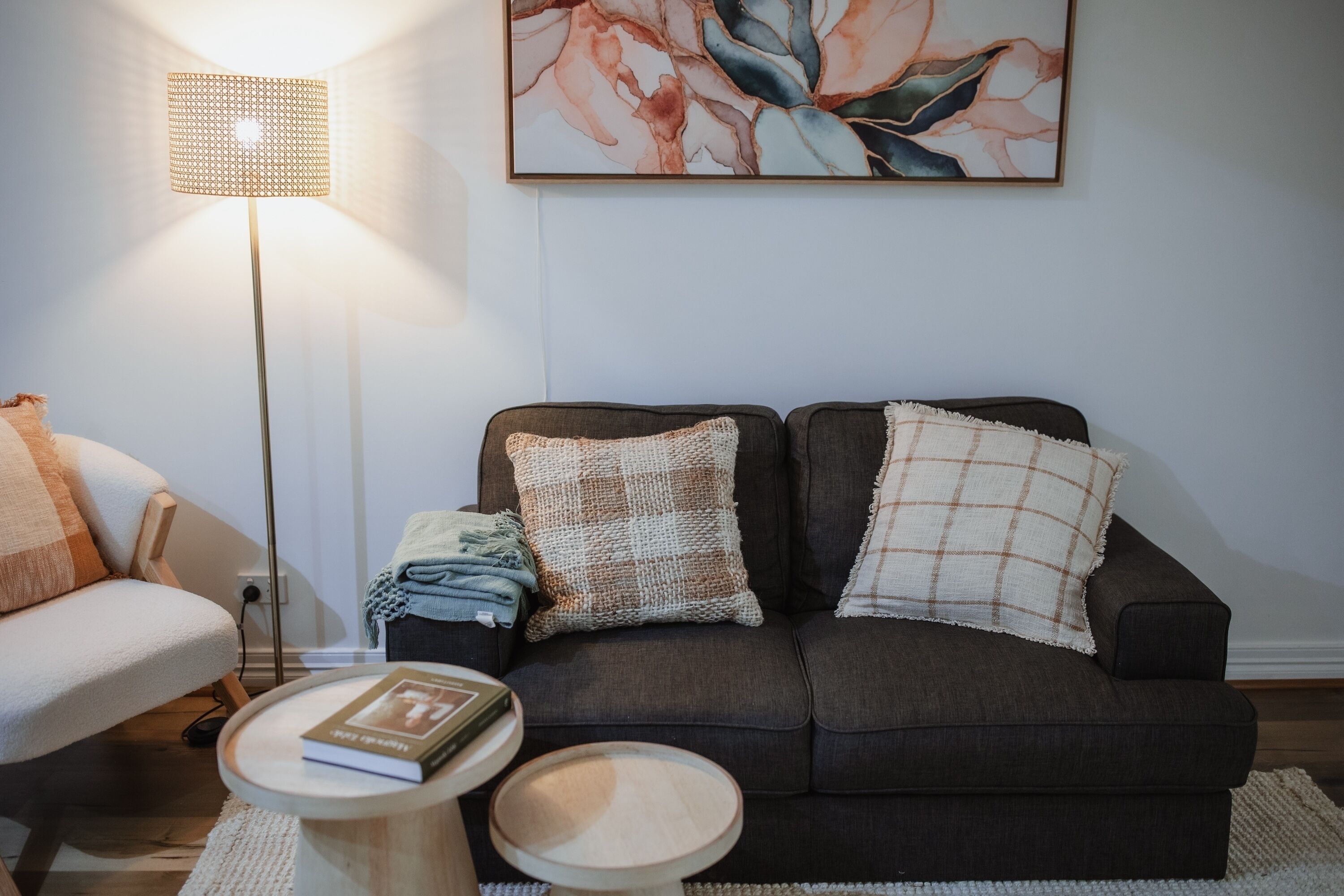 A living room with a dark brown sofa, two pillows, a small round wooden table with a book, a floor lamp, and a painting on the wall.