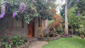 Ein Haus aus Stein mit einer Holztür und -fenstern, umgeben von einem gepflegten Garten mit verschiedenen Pflanzen und einer Hängematte.