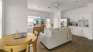 A modern living room with a wooden dining table, a grey sofa, and a kitchen area with white cabinets and appliances. - Beautiful Townhouse IN THE VERY HEART OF DT MT, DORA! (Mount Dora)