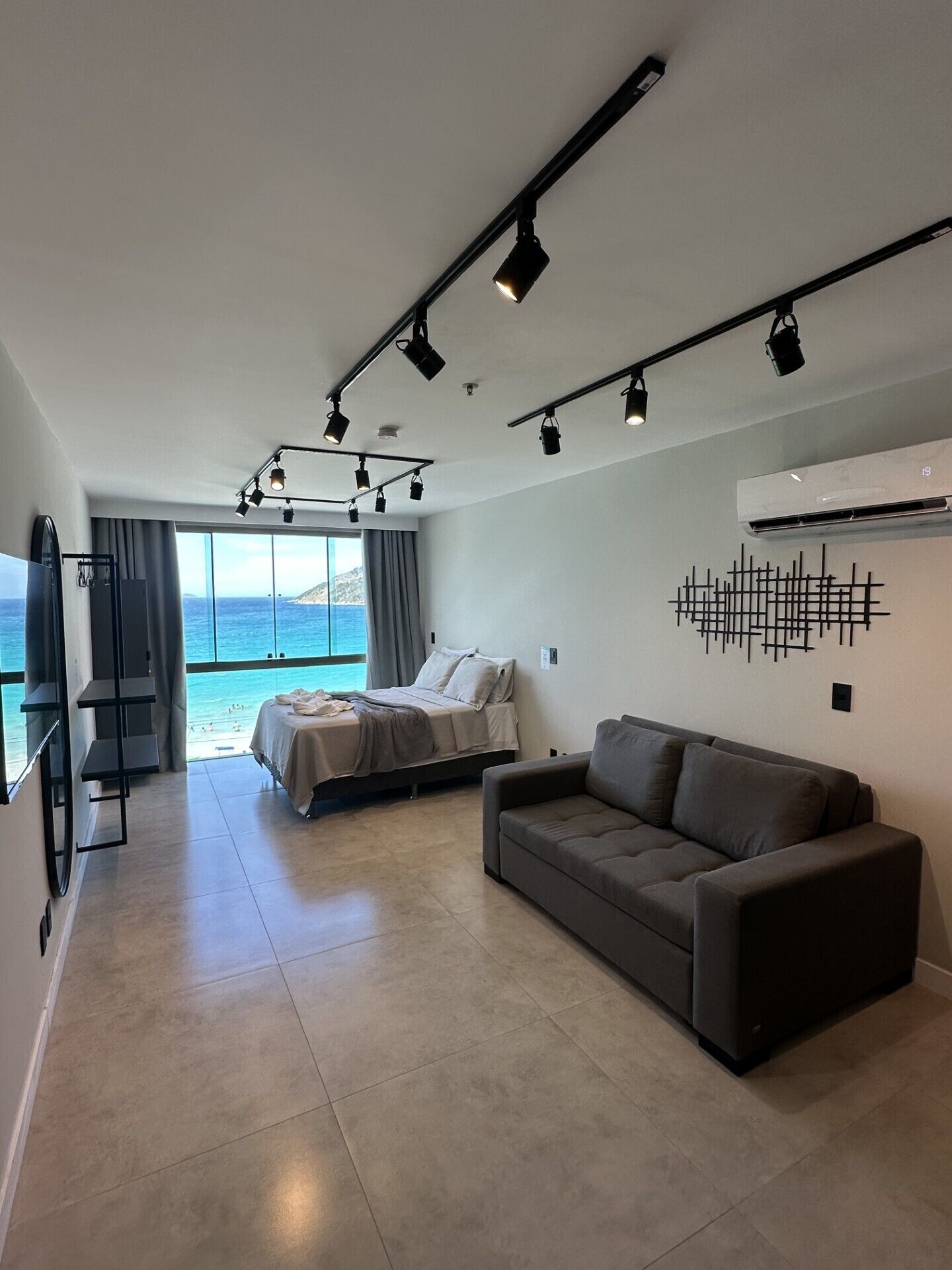 A modern hotel room with a bed, a sofa, a TV, and a large window overlooking the sea.