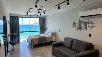 A modern hotel room with a bed, a sofa, a TV, and a large window overlooking the sea.