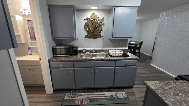 A compact kitchen with grey cabinets, a double sink, and a microwave. There's a decorative wall piece above the sink and a welcome mat at the entrance. - Cozy and Serene 2-bedroom pet friendly house with AC in Downtown Lorain (Lorain)