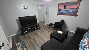A living room with a flat-screen TV, black sofas, a coffee table, a painting of a lighthouse, and a small black chair. - Cozy and Serene 2-bedroom pet friendly house with AC in Downtown Lorain (Lorain)