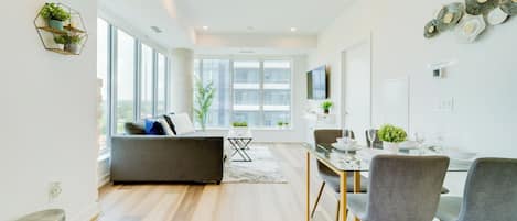 A modern living room with a sofa, dining table, and chairs. There is a large window providing natural light and a view of the city.