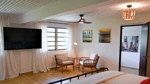Une chambre d’hôtel avec un lit, deux chaises, un téléviseur, un ventilateur de plafond et des œuvres d’art accrochées aux murs.
