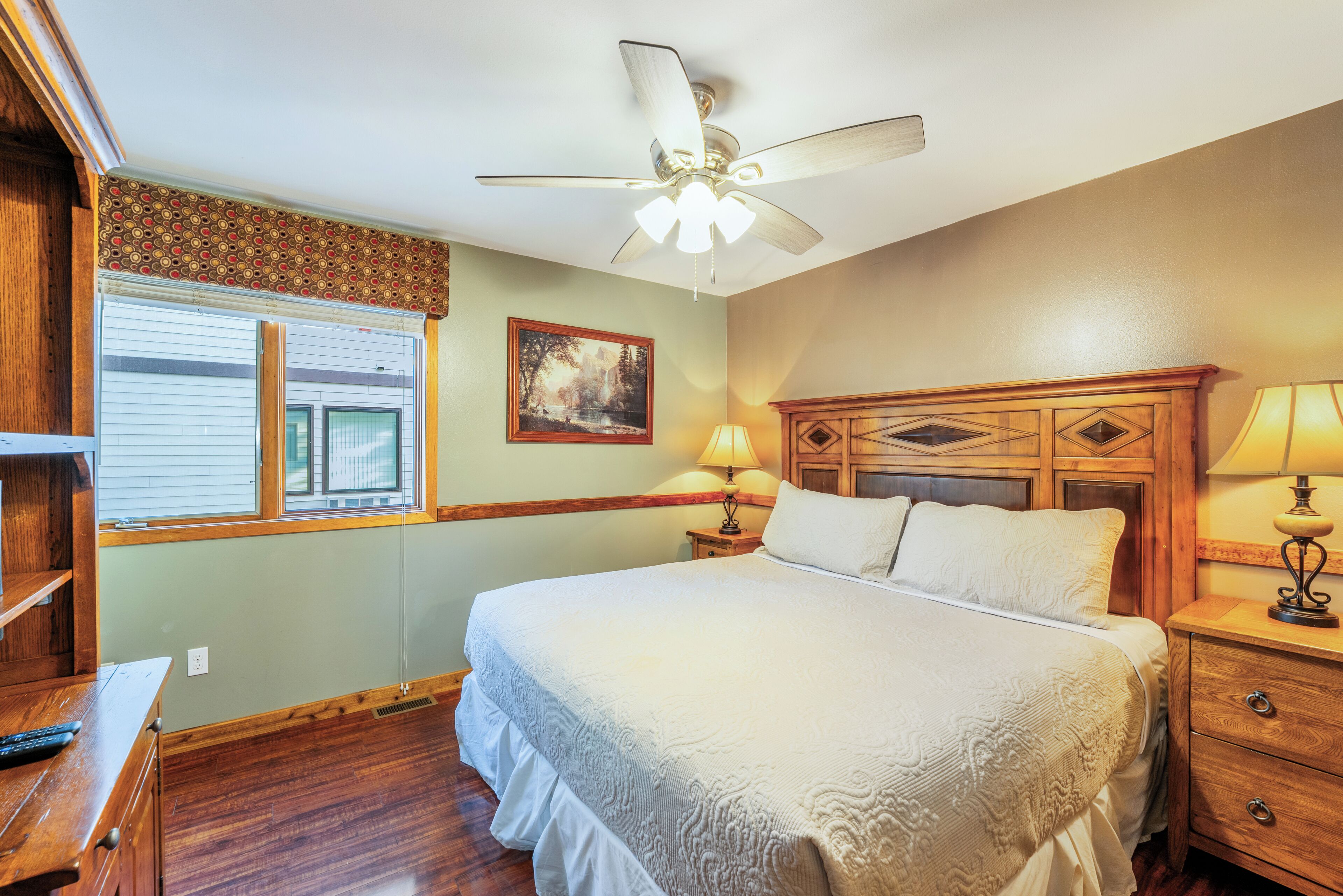 A bedroom with a large bed, wooden headboard, a ceiling fan, a window with blinds, a nightstand with a lamp, and a wooden dresser.