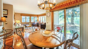 A spacious dining area with a wooden table and chairs, a chandelier, and large windows offering a view of the outdoors.