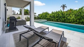 Ett modernt utomhusområde med en pool, solstolar, en grill och utsikt över en frodig, grön häck och en palmtree.