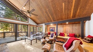 A spacious living area with a large wooden ceiling, leather furniture, and a view of the forest through large windows.