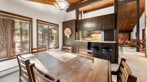 A dining area with a wooden table and chairs, a kitchen with dark cabinets, and large windows with a forest view.