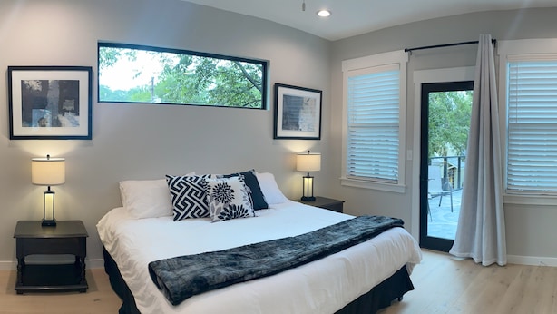 A bedroom with a large bed, two bedside tables with lamps, a view of the outdoors through the window, and a door leading to a balcony.