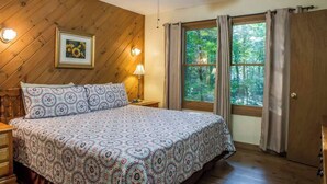 Una camera da letto con soffitto in legno, un letto con biancheria a motivi, un comodino con lampada, un quadro incorniciato al muro e una finestra con tende.