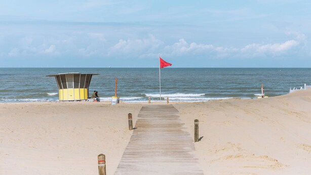 A wooden boardwalk leading to a lifeguard tower and beach area with a red flag.