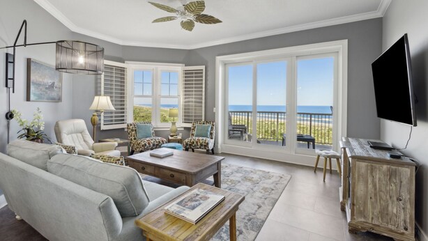 A spacious living room with a large window offering a view of the ocean, a ceiling fan, and a flat-screen TV mounted on the wall. - Ocean Place 22 - Sea Forever (Fernandina Beach)