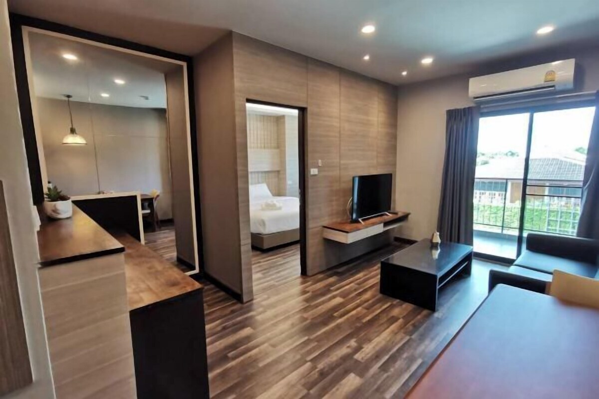 A modern hotel room with a wooden floor, a TV, a sofa, a dining table, and a balcony with curtains.