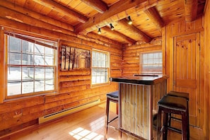 A log cabin interior with a bar area, large windows, and a wooden ceiling.