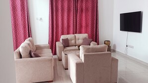 A living room with a beige sofa set, red patterned curtains, a flat-screen TV mounted on the wall, and a small wooden side table. - Sweet Home Ja-Ela (Ja-Ela)
