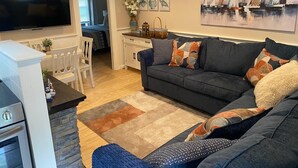 A modern living room with a dark grey sofa, a flat-screen TV, and a kitchen area with a white countertop and wooden cabinets. - Cozy 2-Bed Lakefront Weirs Beach (Laconia)