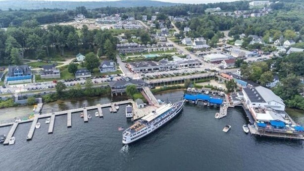 A large cruise ship docked at a pier with a marina, surrounded by residential buildings and greenery. - Cozy 2-Bed Lakefront Weirs Beach (Laconia)