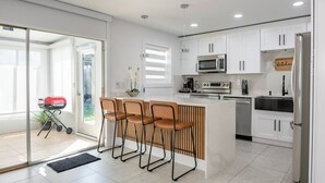 A modern kitchen with white cabinets, a stainless steel refrigerator, a microwave, and a coffee maker. There are three bar stools and a small island with a sink. - Riverside Retreat w Fireplace (Daytona Beach)