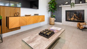 A modern living room with a wooden console, a large potted plant, a fireplace, and a coffee table with books and a board game. - Riverside Retreat w Fireplace (Daytona Beach)