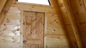Une petite cabane en bois, avec un plafond en pente, un lit simple recouvert d’une couverture rouge et bleue, et une porte en bois.