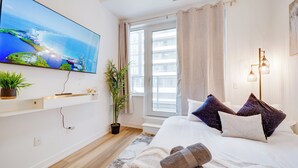 Une chambre d’hôtel moderne avec un grand téléviseur fixé au mur, un lit avec de nombreux oreillers, une plante en pot et une fenêtre avec des rideaux.