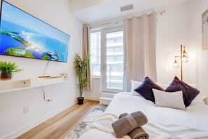 Ein modernes Hotelzimmer mit einem großen Fernseher an der Wand, einem Bett mit vielen Kissen, einer Zimmerpflanze und einem Fenster mit Vorhängen.