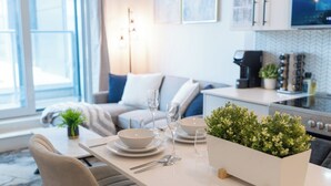 Un espace repas moderne avec une table dressée pour deux personnes, un canapé en arrière-plan et une cuisine visible à travers une porte ouverte.