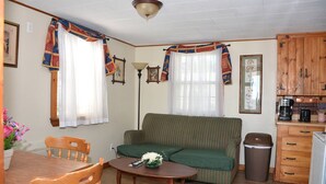 A cozy living room with a green sofa, wooden dining table, and chairs. There is a small kitchen area with wooden cabinets and a trash bin. - Little Miss Cottages - Two Bedroom Cottage (Old Orchard Beach)