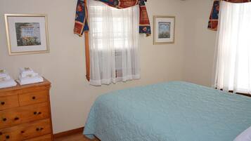 A bedroom with a bed, a wooden dresser, and two windows with sheer curtains and patterned drapes.